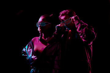 Cool young couple in flashing LED goggles chewing gum and posing in darkness under red light
