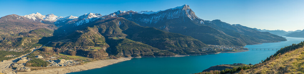 Obraz premium Magnificent panorama of mountains and snow-capped ridges, with a large lake in the valley with turquoise waters and a long road bridge and surrounding villages. Southern Alps, France.
