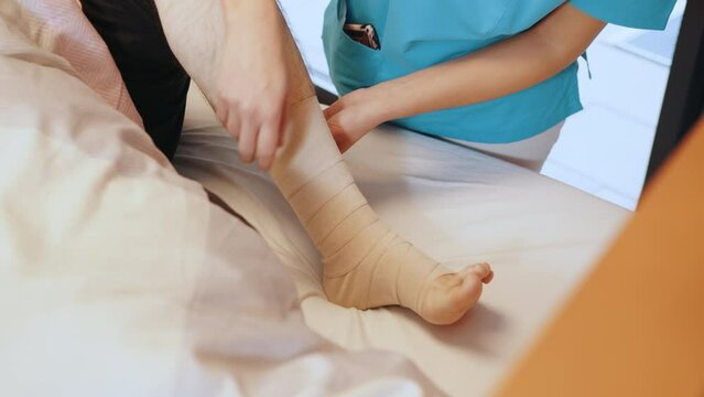 Close-up of male doctor bandaging foot of female patient at doctor's office. Woman Tying Bandage On Patient's Foot. Nurse bandages a patient's leg with an elastic bandage in a hospital bed.