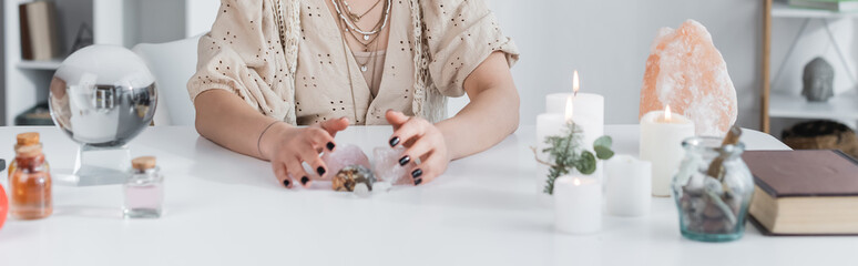 Cropped view of medium touching crystals near candles and magic orb at home, banner.