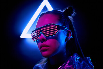 Portrait of serious super girl in neon flashing goggles standing in dark room with neon illumination