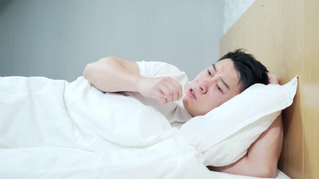 Asian man lying sick in bed with runny nose coughing and flu at home covered with a blanket. Blows her nose into a napkin. Male with fever and headache. Disease. Infection, epidemic, bacillus carrier