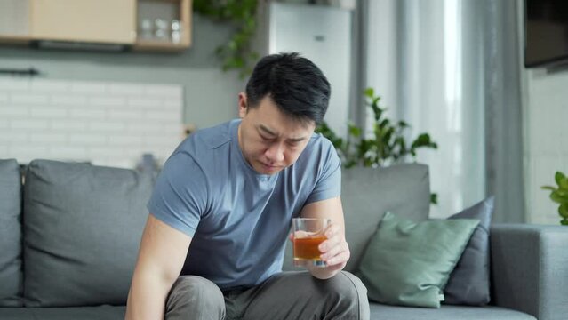 Drunk asian man drinking alcohol with grief sitting at home. Depression, Unhappy frustration, alcohol abuse, addiction, bout set people concept. Depressed stressful guy male alcoholic sits on couch 