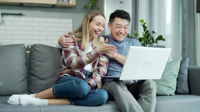 Happy young family wife and husband sit on couch at home check e-mail use laptop read good news online lottery winners receive results, sale discounts, low price excited clients asian couple delighted