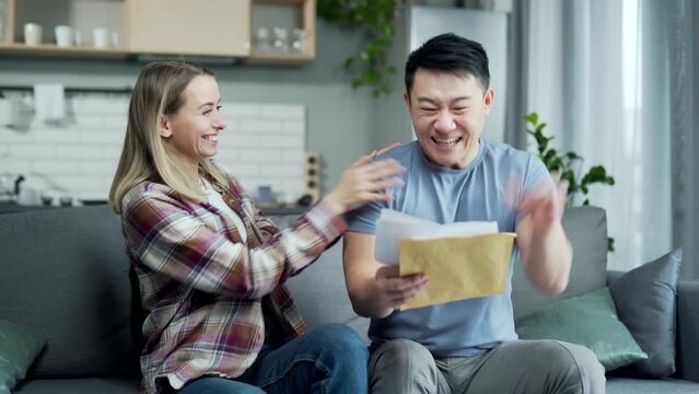 Family receives a letter or message with good news. Happy rejoice. Asian couple member receiving unfolding document rejoicing sitting at home. Cheerful Joyful husband and wife smiling while reading