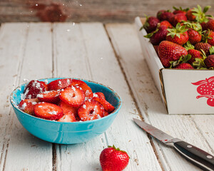 Blue bowl of sliced strawberries, with powdered sugar being sprinkled on them with a carton of strawberries in the background on a white picnic table