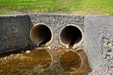 The entrance to the concrete round collector for the river, stream with grass, sand and granite stones on the sides. Hydraulic structures, technologies, hydrology. - obrazy, fototapety, plakaty