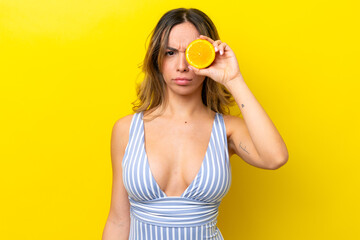 Young caucasian woman isolated on yellow background in swimsuit and holding an orange