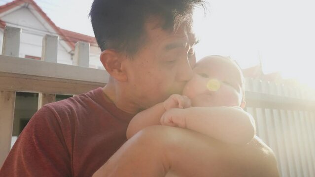 Asian Happy 60s retired old man hugs and kiss on a chubby face of newborn baby in his arms. Close up Grandpa Comforting his nephew against summer warm light, residential village in background 