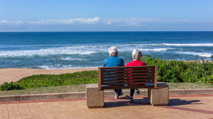 Mature Man Woman Couple Unrecognizable Sitting On Bench Promenade Pathway looking View Of  Beach Ocean Landscape