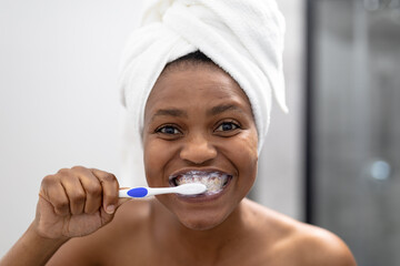 Portrait of african american mid adult woman in towel brushing teeth in bathroom