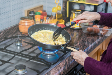Older woman cooks some scrambled eggs in the pan on the gas stove. 