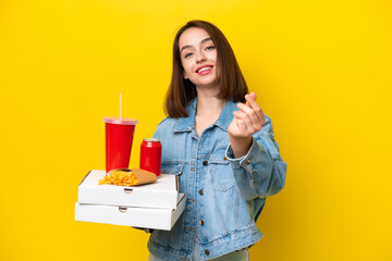 Young Ukrainian woman holding fast food isolated on yellow background making money gesture
