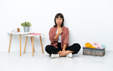 Young mixed race woman folding clothes sitting on the floor isolated on white background surprised and pointing side