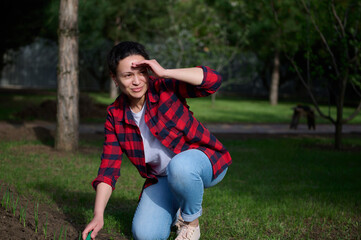 Beautiful serene woman in a checkered shirt, looking smiling aside, covering her face with her hand from the sunlight working on the black soil