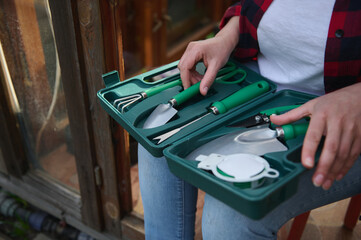 Close-up of a suitcase with a set of garden tools in the hands of a gardener sitting at the entrance to an old wooden greenhouse