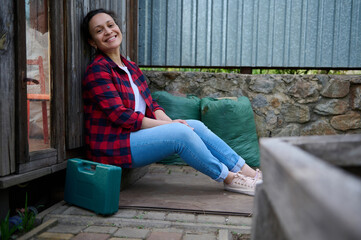 Charming woman gardener sits at the entrance to her wooden greenhouse near suitcase with setof...