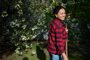 Fototapeta premium Charming middle-aged woman housewife gardener cleaning the local area and raking grass against blossoming fruit tree background on a sunny spring day