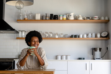 Smiling mid adult african american woman looking away while having coffee in kitchen at home