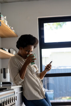 Smiling mid adult african american woman using smart phone while having coffee in kitchen at home