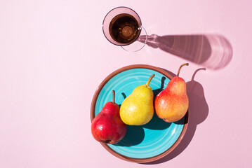 pears of different colors lie on a green plate with a glass and with shadows on a pink background