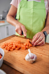 Adult woman, cooking a Spanish dish, estofado de albóndigas. Cutting carrots at the time. Wearing an apron, carrying glasses and cooking utensils.