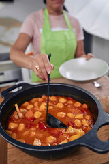 Adult woman, cooking a Spanish dish, estofado de albóndigas. Taking a cell phone photo of the finished meal for social media. Wearing an apron, wearing glasses and cooking utensils.