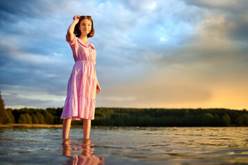 Beautiful teenage girl wearing pink dress having fun by a lake on warm and sunny summer day. Pretty young girl on a sunset.