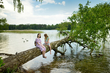 Two cute young girls sitting on a faller tree by the river or lake dipping their feet in the water...