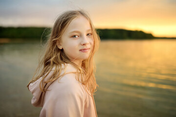Beautiful young girl having fun by a lake on warm and sunny summer day. Pretty child on a sunset.