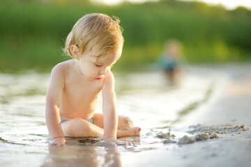Cute toddler boy wearing swimming diaper playing by a river on hot summer day. Adorable child having fun outdoors during summer vacations. - obrazy, fototapety, plakaty