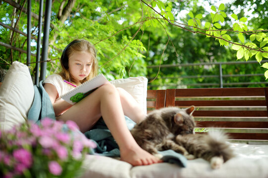 Cute young girl snuggling up on the sofa on a cozy outdoor terrace. Child drawing in a garden under a blanket a on sunny summer evening.