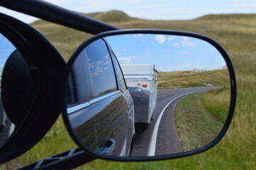 Side rearview mirror reflects side of black vehicle and the pop-up camper being towed, blue skies, long curving road, and grasslands in South Dakota.