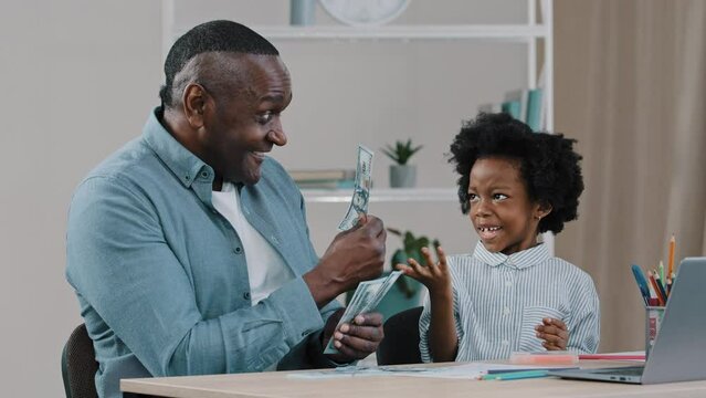 African american father helps to do homework teaches little daughter arithmetic using dollar bills kid girl learns to count dad shows paper money educates math happy family sitting at table home study
