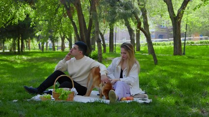 A young man smokes a cigarette on a picnic with his girlfriend, the girl waves her hand from the smoke. A young couple is sitting in the park on a picnic and stroking their dog on the head. Picnic in - Powered by Adobe