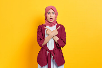 Shocked beautiful Asian woman in casual shirt crossed arms and points sideways, chooses between two options, indicates left and right isolated over yellow background