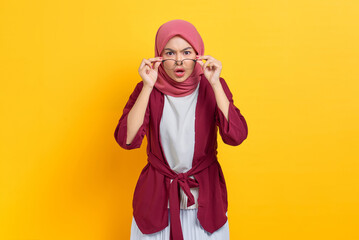 Amazed beautiful Asian woman in casual shirt looking at camera taking off eyeglasses, shocked by news or special offer isolated over yellow background