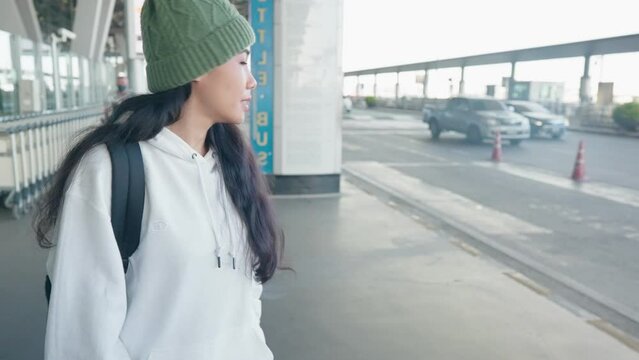 Asian female tourist walking outside a public transportation station or airport terminal hall, waiting for online taxi from application. Young attractive traveler woman walks with suitcase at airport 