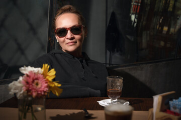 MUJER VESTIDA DE NEGRO CON LENTES OSCUROS, TOMANDO UN CAFE EN UN BAR.