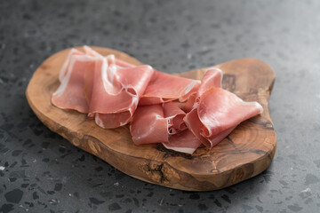 Thin italian prosciutto slices on olive wood board on conctere background
