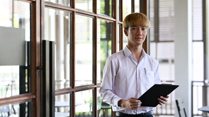 Portrait of confident asian male employee holding clipboard and standing in modern office.