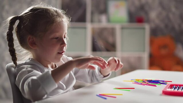 Funny little girl with braids stretches arms and puts tongue out preparing to count colorful sticks at mental arithmetic lesson closeup slow motion