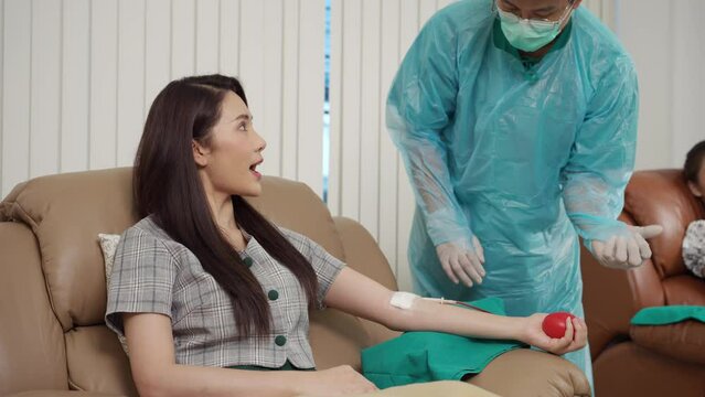 Healthcare worker in protective workwear, CPE Gown suggests the blood donor squeeze the ball for blood to come out easily. Asian woman, 28 years old, Blood Donation and charity concept