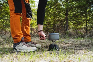 Tourist utensils stand on a gas burner, feet in boots, orange pants, a hand holding a pot of water,...