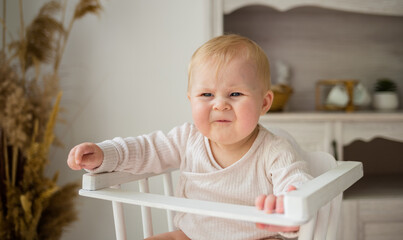 a naughty little girl in a white cotton bodysuit is sitting in a wooden chair in the kitchen