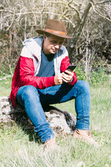 Naklejka na ściany i meble Entrepreneurial self-employed man wearing hat, looking at his mobile while relaxing in the countryside, sitting on a stone. Concept of work, enjoy, relax, Internet, technology and connectivity.