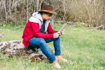Entrepreneurial self-employed man wearing hat, looking at his tablet while relaxing in the field, sitting on a stone. Concept of work, enjoy, relax, Internet, technology and connectivity.
