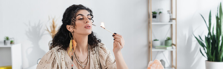 Gypsy fortune teller blowing on burning match near crystal at home, banner.