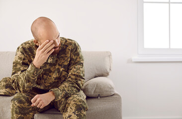 Sad upset depressed veteran soldier suffering from PTSD sitting on couch at home in his military uniform and thinking about horrors of war. Trauma, post traumatic stress disorder concept
