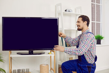 Fototapeta na wymiar Repairman or technician fixing a TV at home. Young man in blue uniform overalls using a screwdriver while repairing, installing or checking a modern large television set in the living room